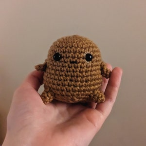 May include: A handmade, brown crocheted creature with black safety eyes and a small smile. The amigurumi figure has small arms and legs, and is held in a hand. The plush toy is a unique, handcrafted item.
