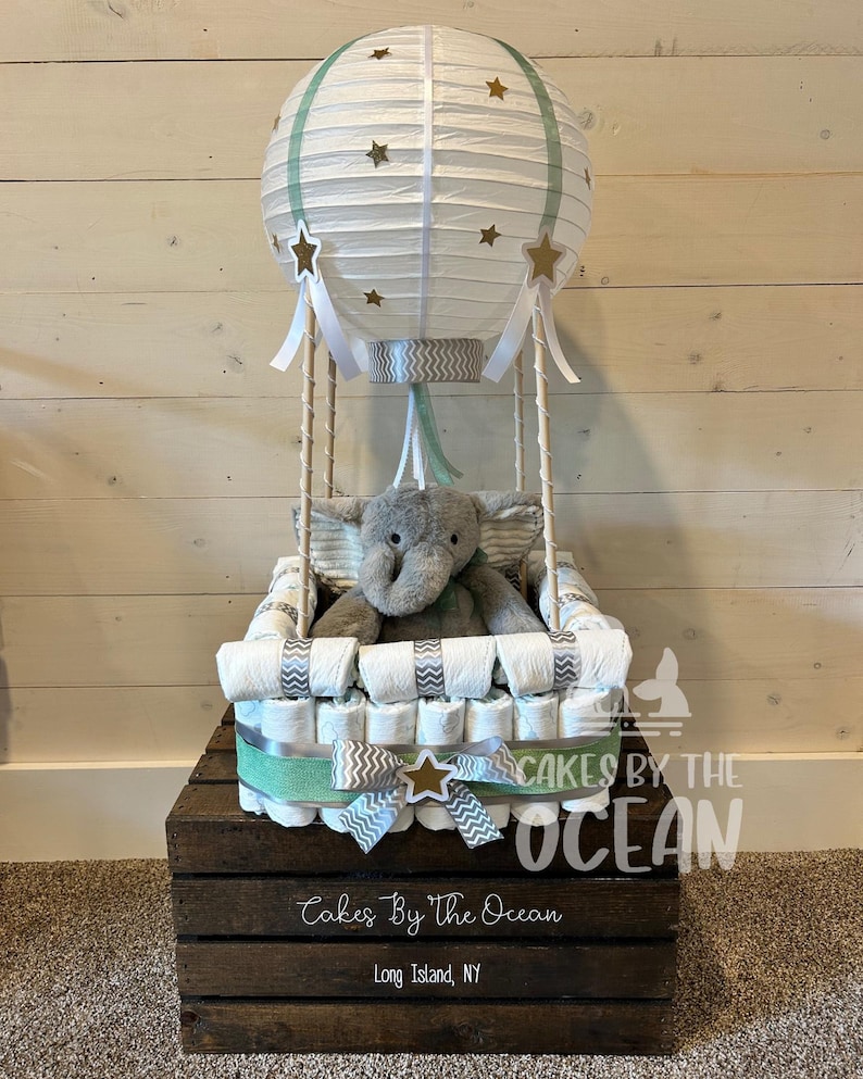 May include: A white hot air balloon diaper cake with a gray elephant plush toy inside. The balloon is decorated with gold stars and green stripes. The cake is sitting on a wooden crate with the text "Cakes By The Ocean" and "Long Island, NY".