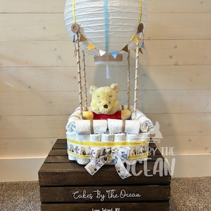 May include: A Winnie the Pooh themed diaper cake in the shape of a hot air balloon. The balloon is made of white paper with yellow and blue stripes and has a brown burlap basket. The cake is decorated with Winnie the Pooh and friends characters.