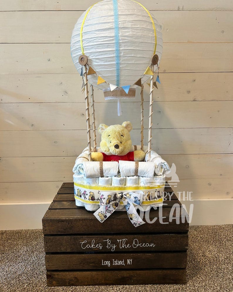 May include: A Winnie the Pooh themed diaper cake in the shape of a hot air balloon. The balloon is made of white paper with yellow and blue stripes and has a brown burlap basket. The cake is decorated with Winnie the Pooh and friends characters.