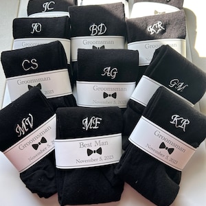 May include: A group of black socks with white embroidered initials and the word "Groomsman" or "Best Man" on white labels. The labels also include the date "November 5, 2023".