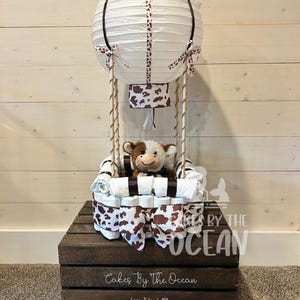 May include: A white and brown diaper cake shaped like a hot air balloon with a stuffed cow inside. The balloon is decorated with brown and white polka dots and a brown ribbon. The cake is sitting on a wooden crate with the text "Cakes By The Ocean" and "Long Island, NY".