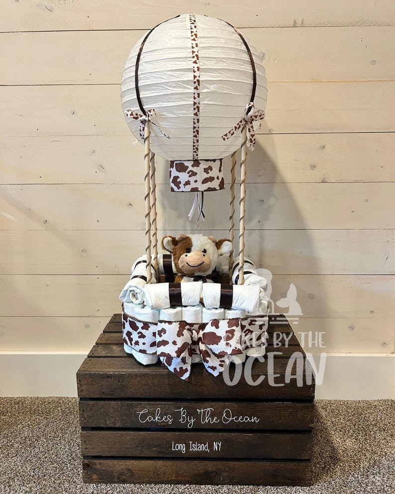 May include: A white and brown diaper cake shaped like a hot air balloon with a stuffed cow inside. The balloon is decorated with brown and white polka dots and a brown ribbon. The cake is sitting on a wooden crate with the text "Cakes By The Ocean" and "Long Island, NY".