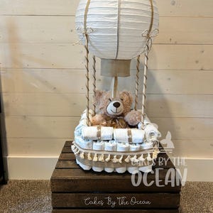 May include: A white hot air balloon diaper cake with a brown teddy bear sitting inside. The balloon is made of paper and has a brown burlap base. The cake is decorated with brown and white ribbon and is sitting on a wooden crate with the text "Cakes By The Ocean" and "Long Island, NY".