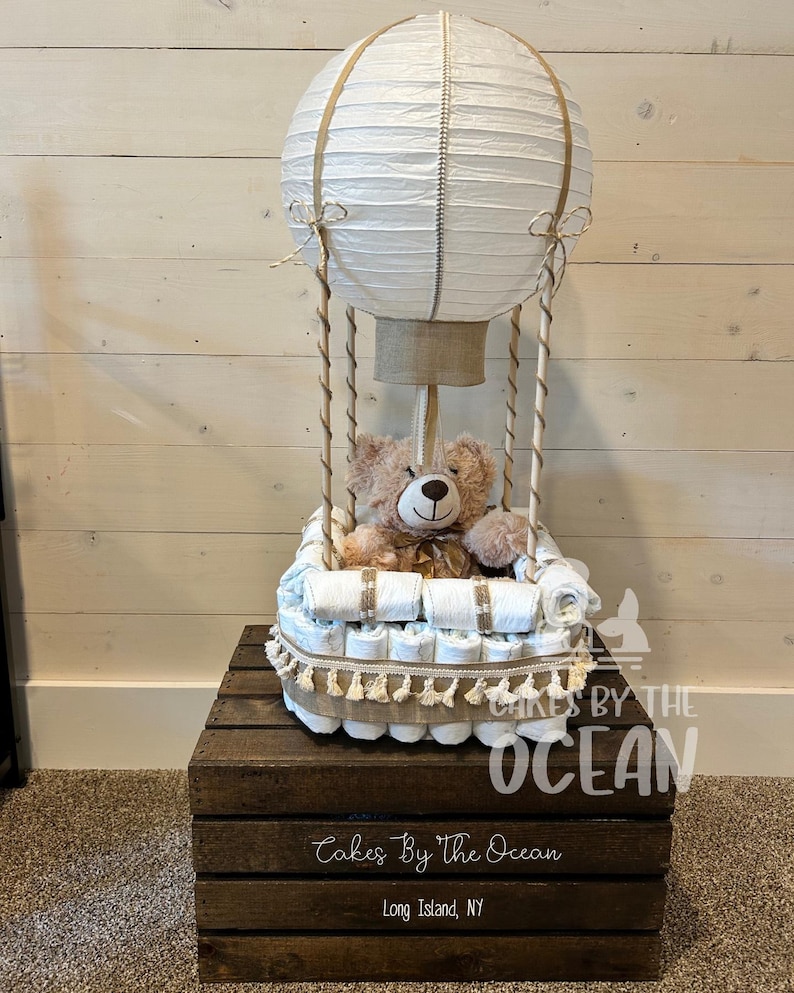 May include: A white hot air balloon diaper cake with a brown teddy bear sitting inside. The balloon is made of paper and has a brown burlap base. The cake is decorated with brown and white ribbon and is sitting on a wooden crate with the text "Cakes By The Ocean" and "Long Island, NY".