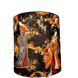 K&ouml;nnte beinhalten: Ein schwarzer Lampenschirm mit einem floralen Muster, das Geishas in traditioneller japanischer Kleidung zeigt. Der Lampenschirm hat einen goldenen Sockel mit drei Beinen.