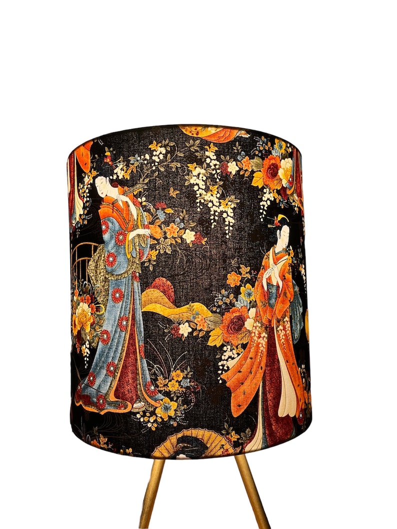 K&ouml;nnte beinhalten: Ein schwarzer Lampenschirm mit einem floralen Muster, das Geishas in traditioneller japanischer Kleidung zeigt. Der Lampenschirm hat einen goldenen Sockel mit drei Beinen.