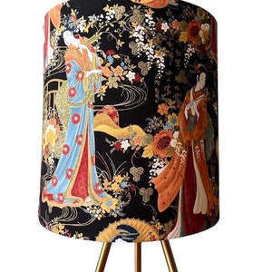 K&ouml;nnte beinhalten: Ein schwarzer Lampenschirm mit einem goldenen und floralen Muster, das japanische Frauen in traditioneller Kleidung zeigt. Der Lampenschirm steht auf einem goldenen Dreibein-St&auml;nder.