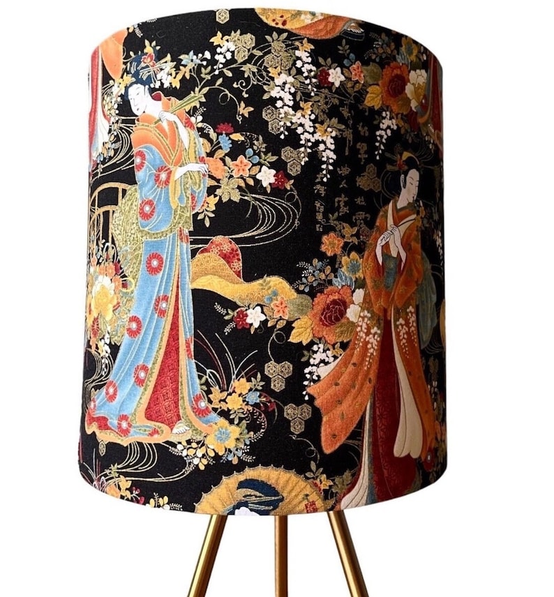 K&ouml;nnte beinhalten: Ein schwarzer Lampenschirm mit einem goldenen und floralen Muster, das japanische Frauen in traditioneller Kleidung zeigt. Der Lampenschirm steht auf einem goldenen Dreibein-St&auml;nder.