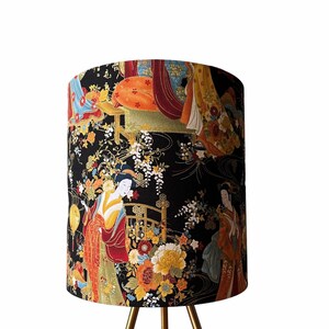 K&ouml;nnte beinhalten: Ein schwarzer Lampenschirm mit einem goldenen Dreibein-St&auml;nder. Der Lampenschirm zeigt einen farbenfrohen japanisch inspirierten Druck mit Geishas und floralen Mustern.