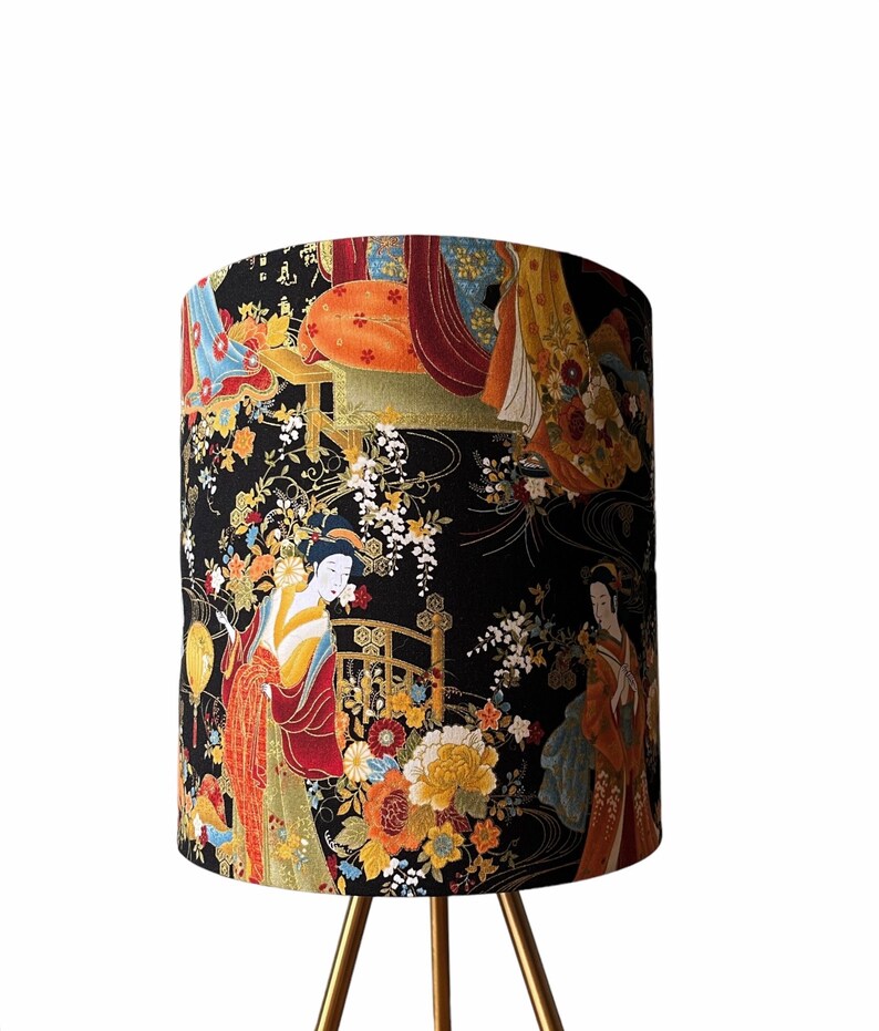 K&ouml;nnte beinhalten: Ein schwarzer Lampenschirm mit einem goldenen Dreibein-St&auml;nder. Der Lampenschirm zeigt einen farbenfrohen japanisch inspirierten Druck mit Geishas und floralen Mustern.