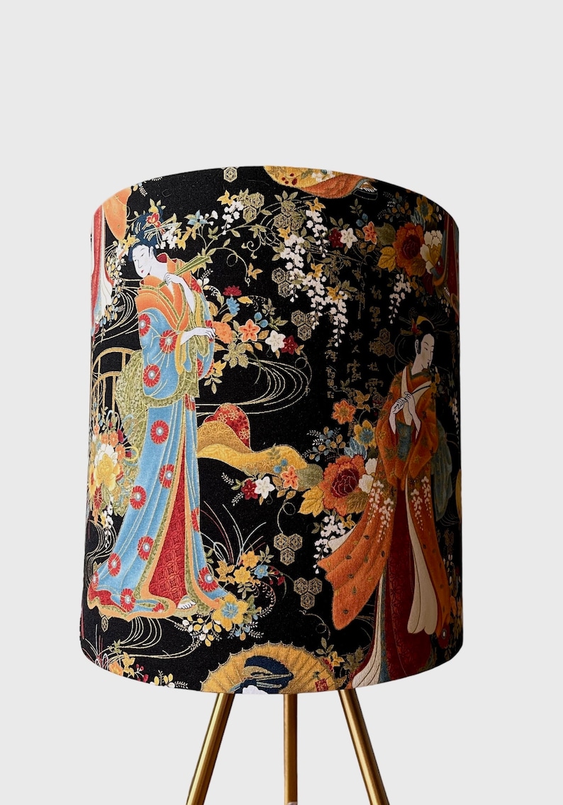 K&ouml;nnte beinhalten: Ein schwarzer Lampenschirm mit einem goldenen Dreibeinst&auml;nder zeigt einen Blumendruck mit zwei Frauen in traditioneller japanischer Kleidung. Der Druck enth&auml;lt goldene Akzente und einen schwarzen Hintergrund.