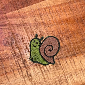 May include: A green and brown embroidered snail patch with a smiling face.