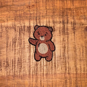 May include: A brown teddy bear patch with a light tan belly and outstretched arm. The patch is embroidered with a textured appearance and a black border. The bear is set against a wooden background.