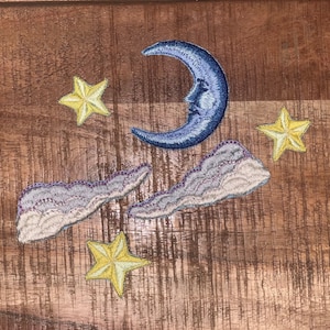 May include: A blue crescent moon with a face embroidered on a wooden surface with three yellow stars and two white clouds with purple accents.