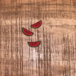 May include: Three embroidered watermelon slice patches. Each patch features a red watermelon slice with a green rind and black seeds. The patches are arranged on a textured, brown wooden surface.