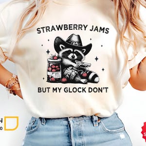 May include: Cream-coloured t-shirt with a raccoon in a cowboy hat holding a gun, and a jar of strawberry jam. The text says "STRAWBERRY JAMS BUT MY GLOCK DON'T".
