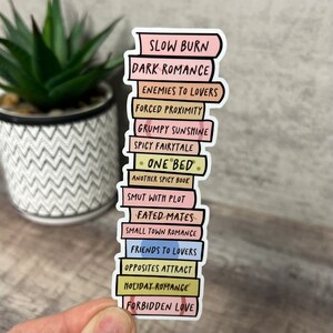 May include: A sticker depicting a stack of books with titles such as "Slow Burn" and "Holiday Romance." The books have pastel-coloured spines and black lettering. The sticker is held against a blurred background.