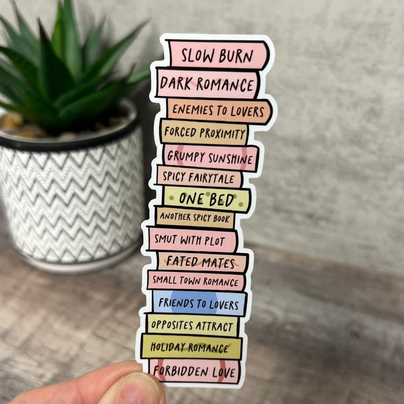 May include: A sticker depicting a stack of books with titles such as "Slow Burn" and "Holiday Romance." The books have pastel-coloured spines and black lettering. The sticker is held against a blurred background.