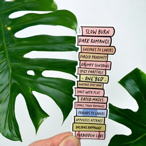 May include: A sticker depicting a stack of books with titles including "Slow Burn" and "Dark Romance." The books are in pastel shades with black text. The sticker is held against a backdrop of a large green leaf, creating a contrasting visual.