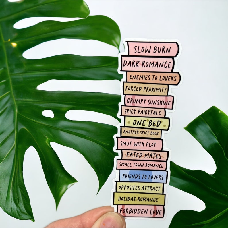 May include: A sticker depicting a stack of books with titles including "Slow Burn" and "Dark Romance." The books are in pastel shades with black text. The sticker is held against a backdrop of a large green leaf, creating a contrasting visual.