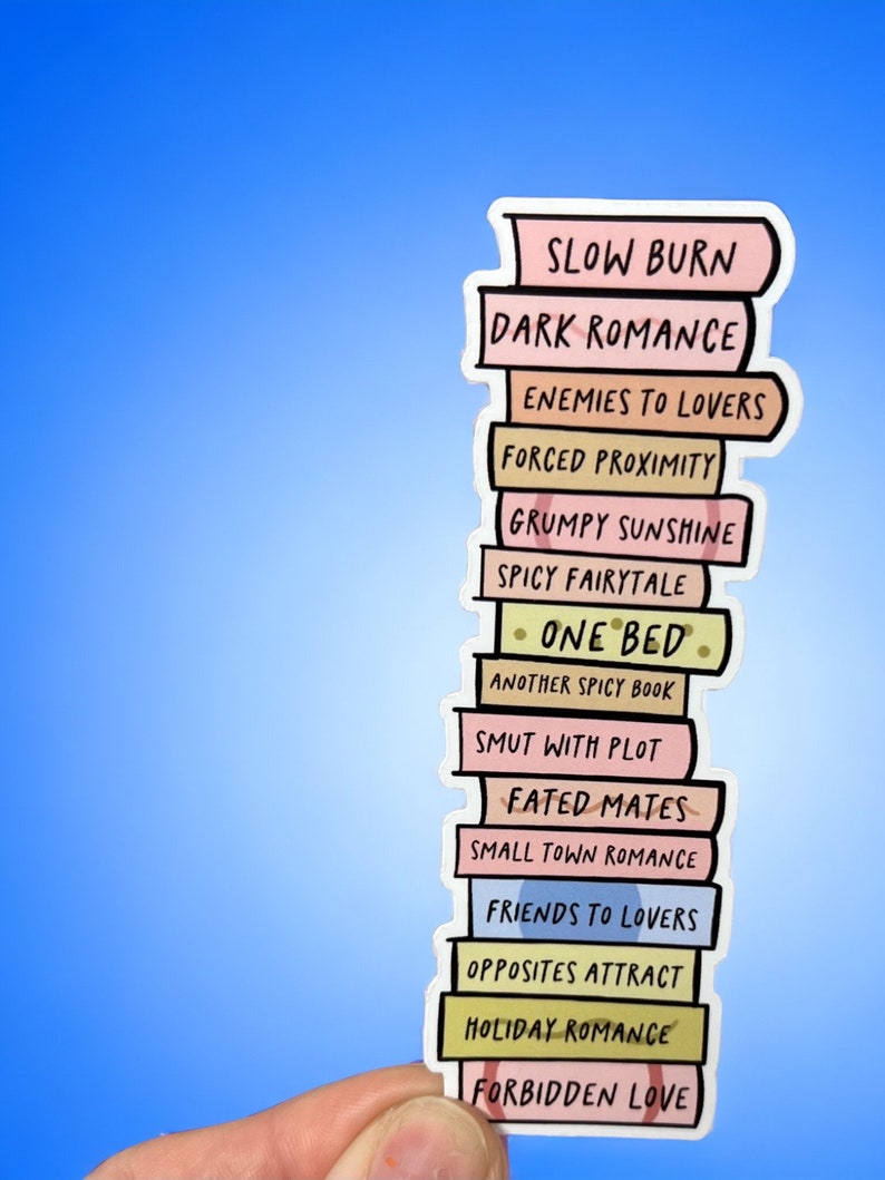 May include: A sticker depicting a stack of books with titles such as "Slow Burn" and "Dark Romance." The books are in pastel shades, held against a blue backdrop.