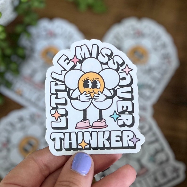 May include: A white sticker with a daisy design and the words "Little Miss Overthinker" in a retro font. The daisy has a yellow center, white petals, and black outlines. The sticker has a black border and star accents.