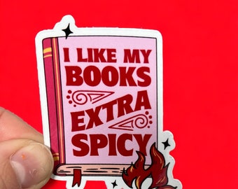 May include: A sticker featuring a pink book with the text "I LIKE MY BOOKS EXTRA SPICY" in red. The book has a red spine and a red ribbon bookmark. Flames and stars are on the bottom right and top corners.