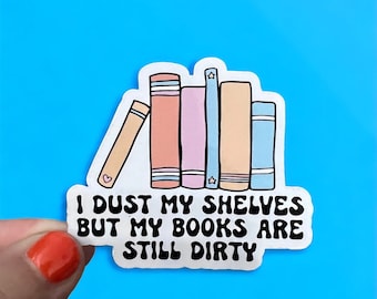 May include: A sticker featuring a row of colorful books with the text "I dust my shelves but my books are still dirty." The books are in shades of peach, pink, blue, and tan. A small heart and stars are on the books.
