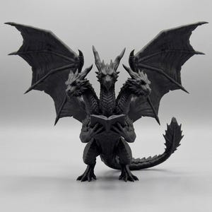 May include: A matte black figurine of a three-headed dragon with large, spread wings. The creature stands on two legs, holding an open book in its front claws. Each head features sharp horns and detailed scales, with a long, spiked tail curving behind.