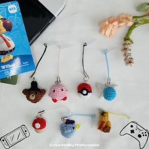 May include: A collection of handmade crochet charms in various shapes, including a bear, a pink creature, a Pokeball, a blue winged creature, a mushroom, a blue bird, and a yellow creature. Each charm has a silver-colored clasp and a black or colored cord.