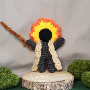 May include: A handmade crochet doll with a black head, gray body, and a flame-like halo of yellow, orange, and red yarn. It wears a cream-colored shawl and holds a brown pipe cleaner wand. The doll is displayed on a wooden slice.