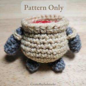May include: A crocheted doll in a pot shape, with a tan body, gray arms and legs, and a red interior. The text "Pattern Only" is at the top of the image. The doll is handmade and sits on a wooden surface.