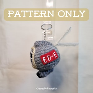 Puede incluir: Un llavero gris tejido a ganchillo con forma de robot, con un parche rojo que dice "ED-E". El llavero tiene una cadena plateada y una pequeña boquilla negra. El texto "PATTERN ONLY" está en la parte superior.