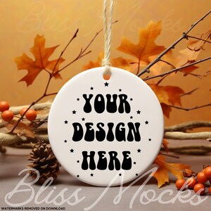May include: A white ceramic ornament with the text "YOUR DESIGN HERE" in black, surrounded by small black stars. The ornament hangs from a natural twine string. Autumn leaves and berries are in the background.