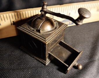 May include: A vintage-style metal pencil sharpener shaped like a coffee grinder. The sharpener has a drawer that pulls out to collect the pencil shavings. The sharpener is made of a dark metal with a worn finish.