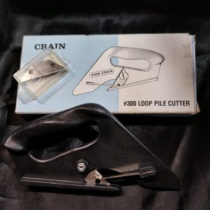 May include: Black loop pile cutter with a curved handle and a silver blade. The box reads "CRAIN" and "#300 LOOP PILE CUTTER". A clear plastic case with extra blades is also visible.