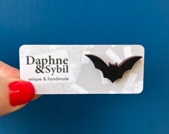 Könnte beinhalten: Eine schwarze fledermausförmige Anstecknadel auf einer rechteckigen weißen Präsentationskarte. Die Karte zeigt den schwarzen Text Daphne & Sybil sowie den Schriftzug unique & handmade. Die Fledermaus hat spitze Ohren und ausgebreitete Flügel.