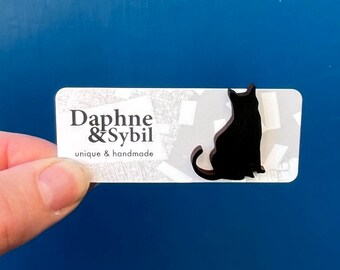 Könnte beinhalten: Eine schwarze, katzenförmige Anstecknadel auf einer weißen, rechteckigen Präsentationskarte. Die Karte zeigt den schwarzen Schriftzug "Daphne & Sybil" sowie die Wörter "unique & handmade" in kleinerer Schrift auf grauem Muster.