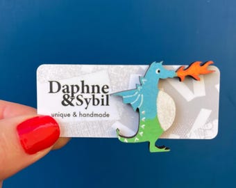 Könnte beinhalten: Eine hölzerne Drachenbrosche mit blauem Kopf und Flügeln, grünem Schwanz und weißem Bauch, die orangefarbene Flammen speit. Die Brosche ist auf einer weißen, rechteckigen Karte mit dem Text Daphne & Sybil unique & handmade befestigt.