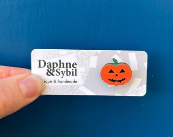 Könnte beinhalten: Eine orangefarbene Kürbis-Anstecknadel mit grünem Stiel und schwarzen, geschnitzten Gesichtszügen. Die Anstecknadel ist auf einer weißen Karte befestigt, die den Text Daphne & Sybil unique & handmade in schwarzer Schrift auf grauem Grund zeigt.