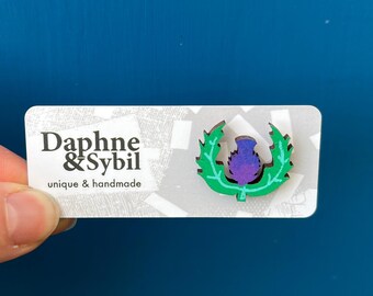Könnte beinhalten: Eine kleine Holzanstecknadel in Form einer violetten und blauen Distel mit grünen Blättern, befestigt auf einer weißen rechteckigen Präsentationskarte. Die Karte zeigt den schwarzen Text Daphne & Sybil und den Schriftzug unique & handmade.