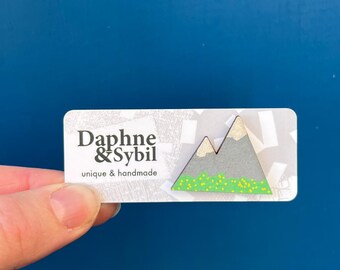 Könnte beinhalten: Eine bergförmige Anstecknadel mit grauen Gipfeln, goldenen Spitzen und einer grünen Basis mit gelben Punkten. Die Nadel ist auf einer weißen, rechteckigen Karte befestigt, die mit dem Text Daphne & Sybil unique & handmade bedruckt ist.