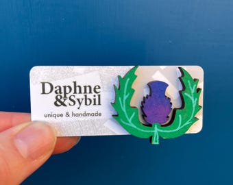 Könnte beinhalten: Eine rechteckige weiße Präsentationskarte mit dem schwarzen Text Daphne & Sybil unique & handmade. Daran befestigt ist eine hölzerne Brosche in Form einer violetten Distelblüte mit hellgrünen Blättern, die dunkle Umrisse und Details aufweist.