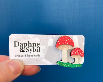 Könnte beinhalten: Eine Holzanstecknadel mit zwei roten Pilzen mit weißen Punkten auf hellem Stiel, befestigt auf einer weißen Karte mit dem Text Daphne & Sybil unique & handmade. Die Pilze stehen auf einer grünen Basis mit kleinen gelben Punkten.