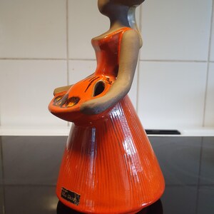 May include: A ceramic figurine of a woman in a long orange dress and hat, holding a basket. The figure has a dark gray face and arms. The dress has vertical lines. The base of the figurine has a label that reads "Ulla Skogh".