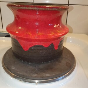 Op de afbeelding: Een handgemaakte keramische vaas met een opvallend tweekleurig ontwerp. Het bovenste gedeelte is helderrood, met een glanzende afwerking en een druppeleffect, in contrast met de donkerbruine basis. De vaas staat op een cirkelvormige, metalen basis.