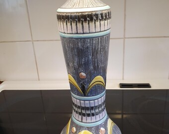 Puede incluir: Un jarrón de cerámica gris alto con un diseño geométrico en tonos de azul, amarillo y blanco. El jarrón tiene una base ancha y un cuello estrecho.