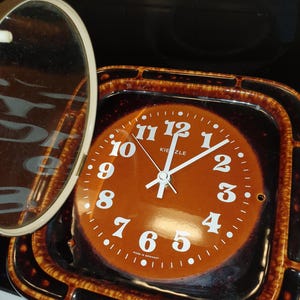 Peut inclure: Une horloge murale carrée Kienzle avec un cadre en céramique marron doté d'une bordure tachetée brun foncé. Le cadran est marron clair avec des chiffres arabes blancs, des aiguilles blanches et l'inscription Kienzle et Made in Germany.