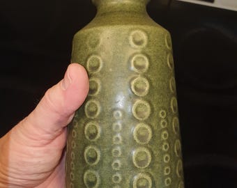 Può includere: Un vaso di ceramica verde con una superficie strutturata con un motivo di cerchi in rilievo. Il vaso ha un corpo cilindrico e un collo leggermente svasato. Il colore è un verde oliva tenue.